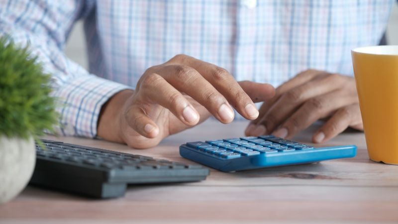 Hands working a blue desk calculator beside a keyboard and a coffee mug
