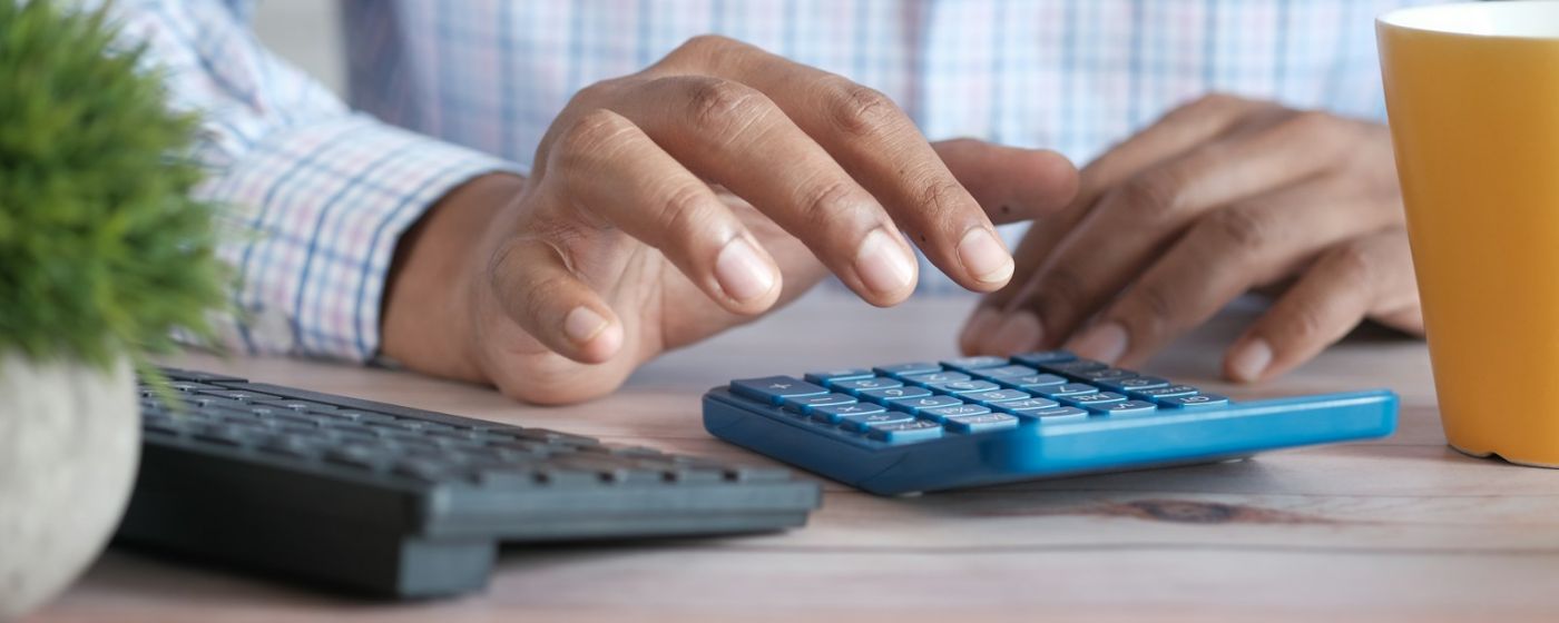 Hands working a blue desk calculator beside a keyboard and a coffee mug