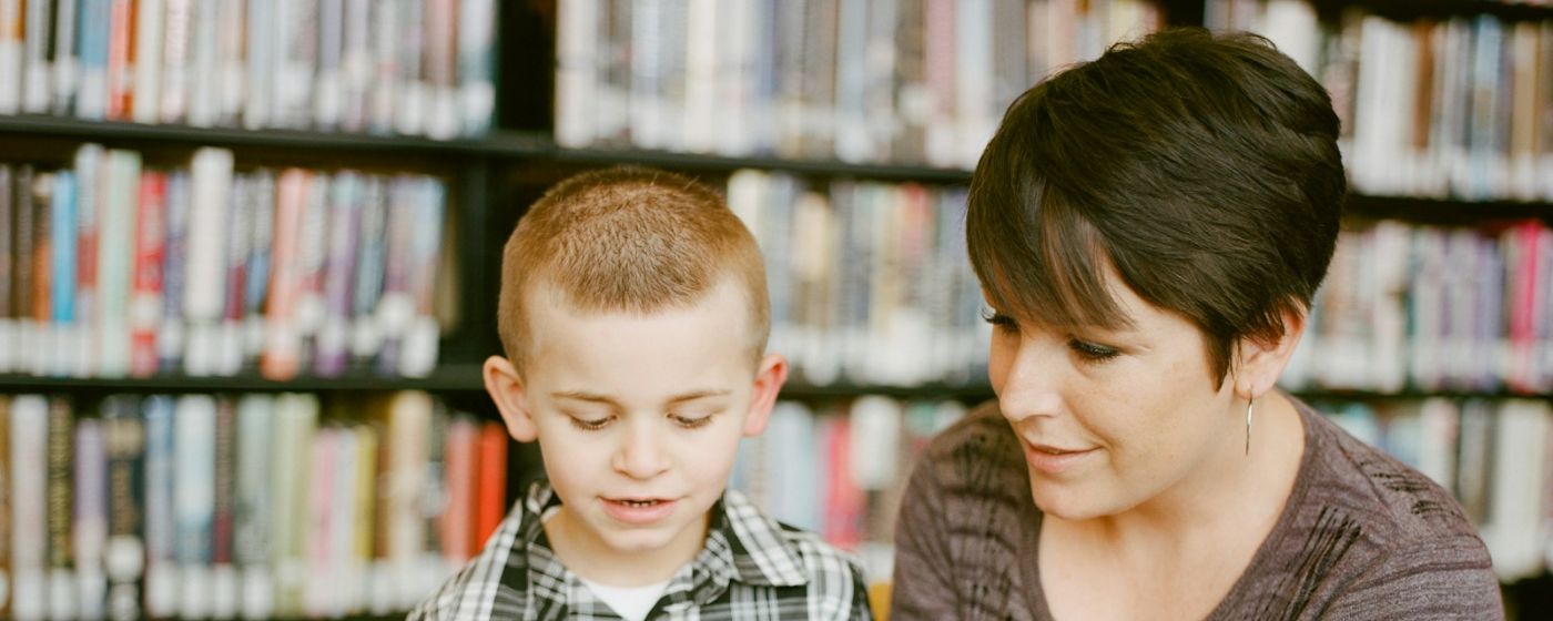 A woman and a young boy read a book together in front of library shelves