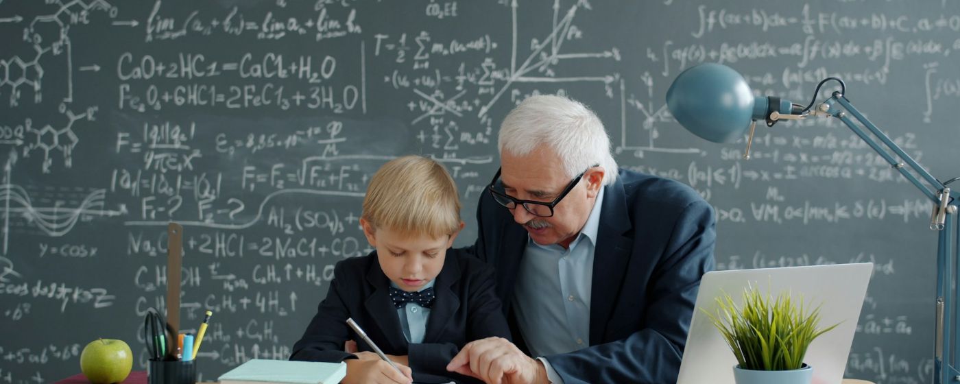 A white-haired teacher helps a young student with a math problem at a desk in front of a chalkboard