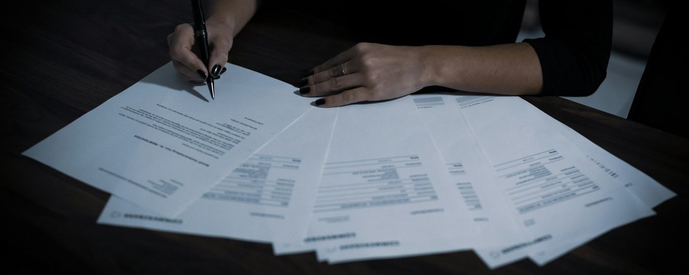 A person marks up a contract with a pen, with pages fanned out across a dark wooden table